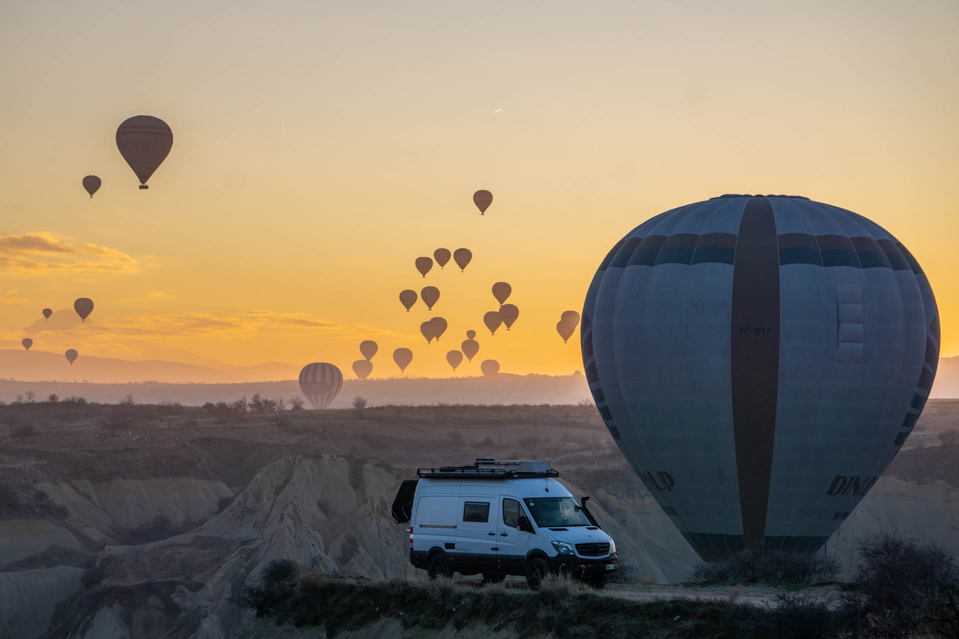Furgoneta camperitzada aparcada en un turó a l'alba, amb globus aerostàtics sobre la vall